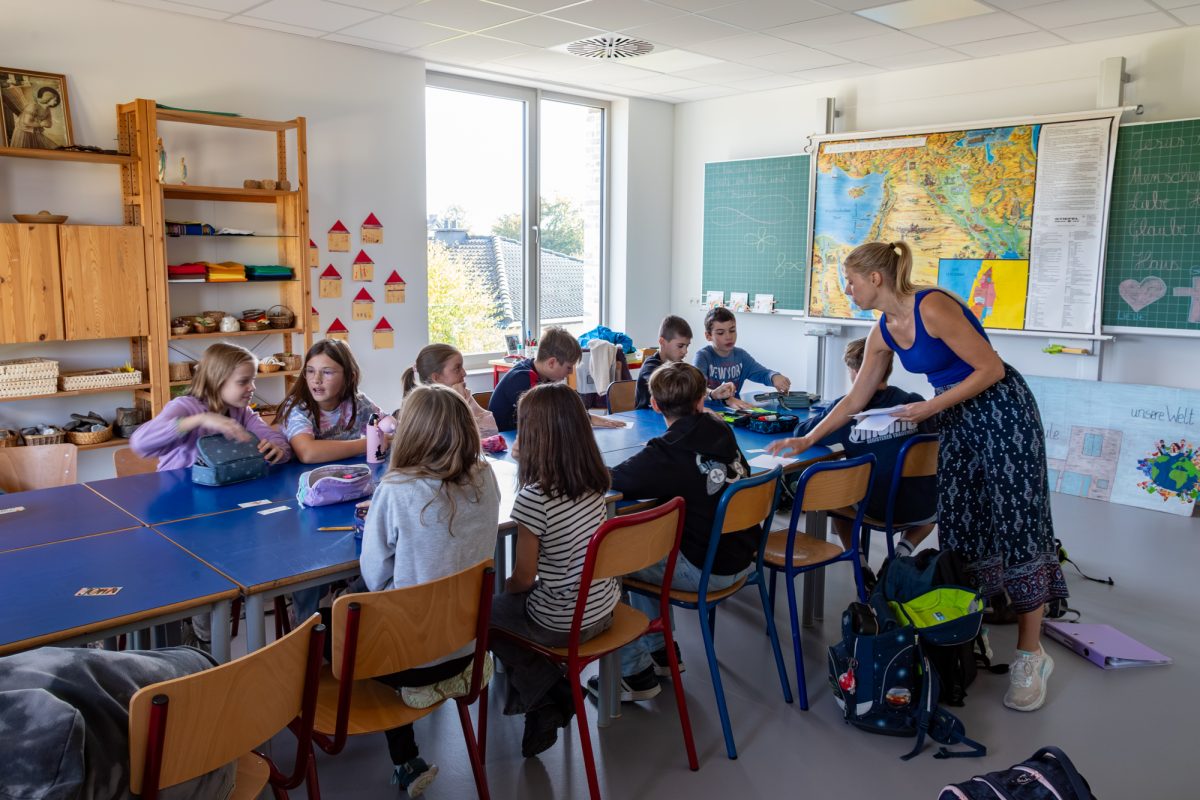 Kinder sitzen an Tischen in einem Klassenraum, eine Lehrerin steht bei ihnen, vor der Tafel hängt eine große Landkarte