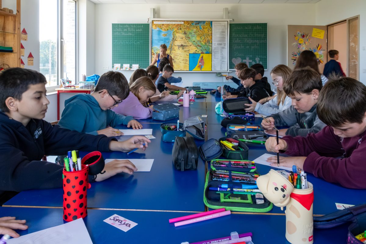 Kinder sitzen an Tischen in Klassenraum und schreiben oder zeichnen etwas, eine Lehrerin befindet sich weiter hinten vor der Tafel - eine Landkarte wurde dort angebracht