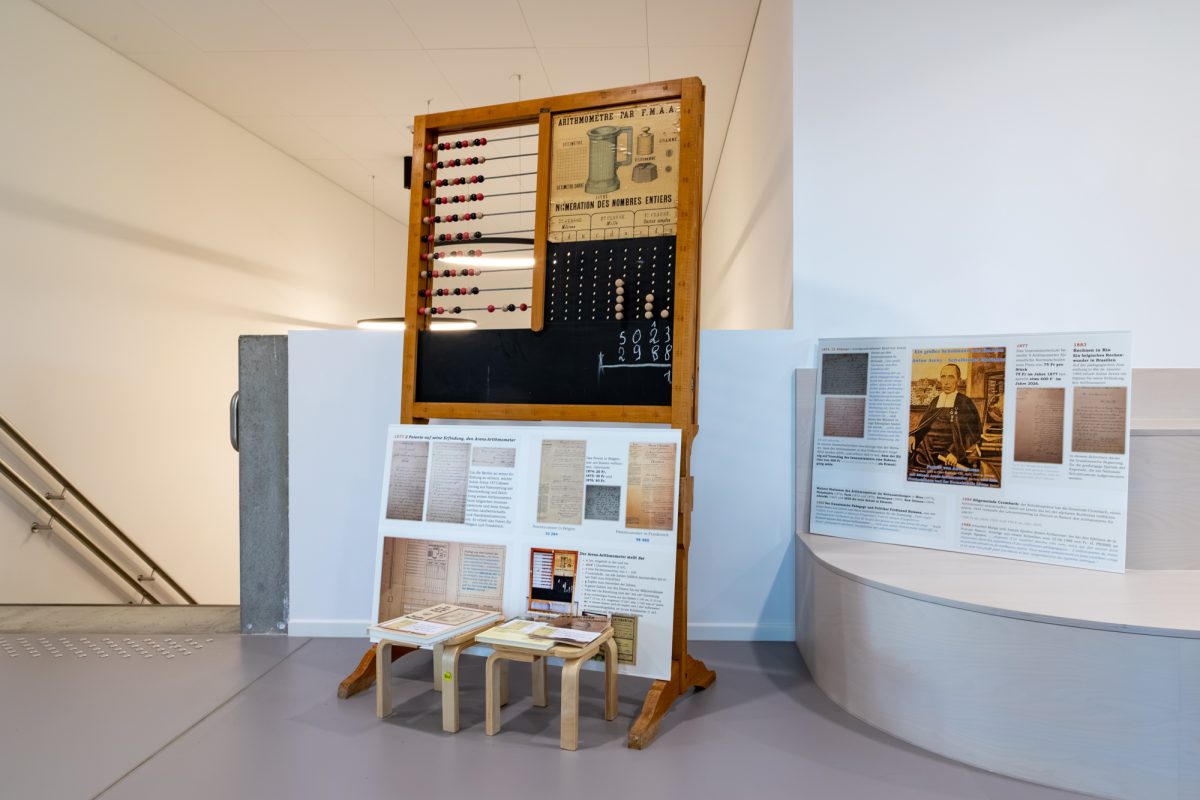 Schulflur, links führt eine Treppe nach unten, in der Mitte stehen das historische Arithmometer und daneben Infotafeln