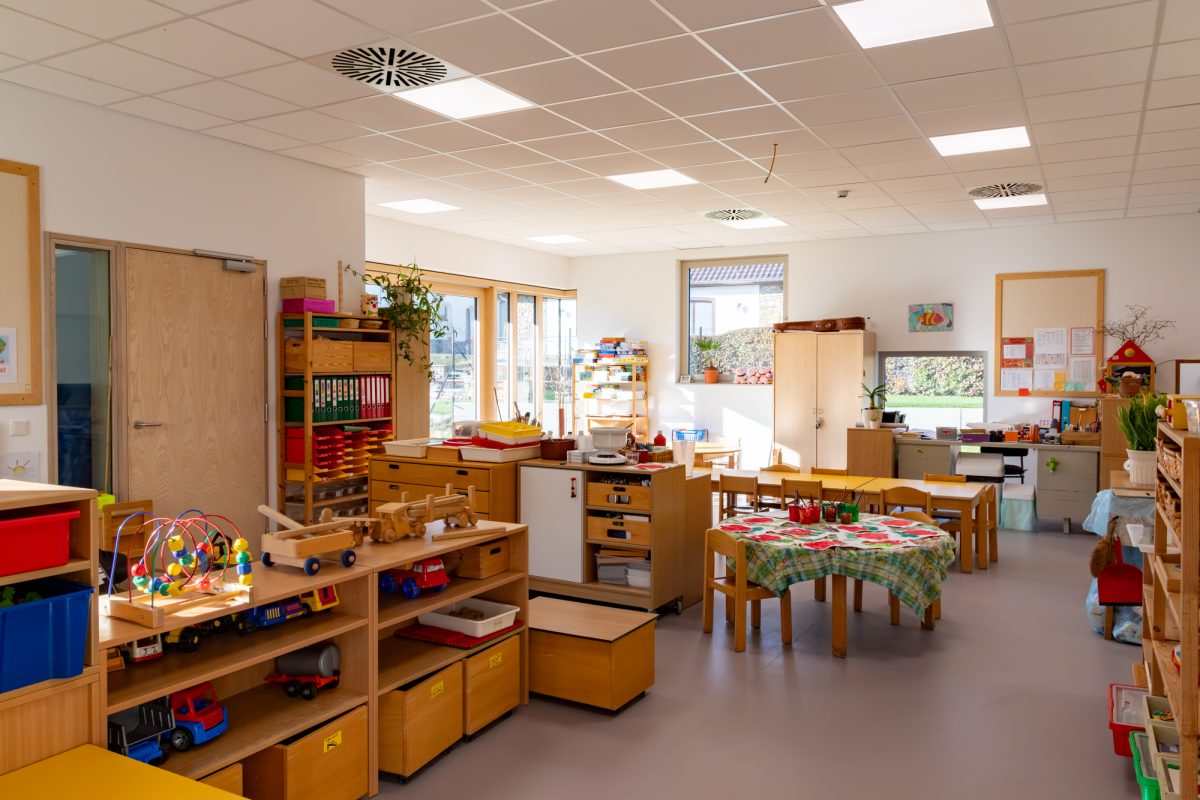 Klassenraum mit Tischen und Stühlen für Kindergartenkinder, außerdem stehen Spielsachen auf Regalen