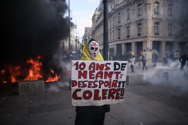 Ein vermummter Demonstrant hält ein Schild mit der Aufschrift "10 Jahre nichts - Verzweiflung und Wut". Im Hintergrund Flammen und Rauchgasschwaden