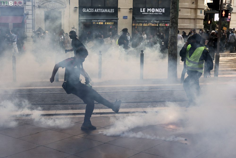 Ein vermummter Demonstrant kickt eine rauchende Tränengaskartusche weg, im Hintergrund sind weitere Demonstranten zu sehen