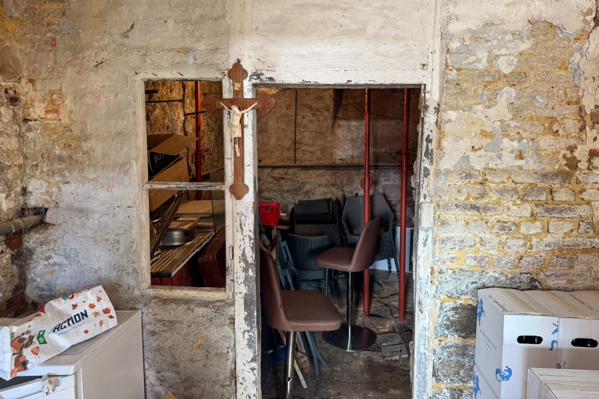 Zugang zum Keller der Vinothek, in dem mehrer Stühle und eine Spüle lagern - ein Kreuz hängt an der Wand