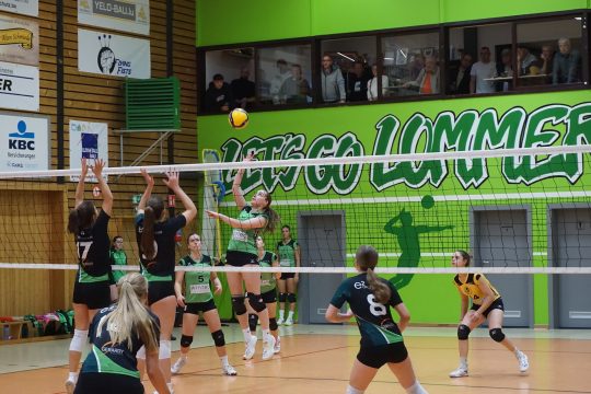 ein Volleyball über dem Netz, Spielerinnen springen auf beiden Seiten hoch